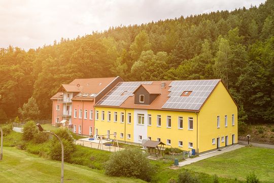 Außenansicht auf das Suchthilfezentrum für Mutter und Kind in Wolfersdorf. Direkt hinter dem gelb und rot gestrichenen Gebäude befindet sich grüner Mischwald. Die Außenanlagen sind sehr gepflegt angelegt.