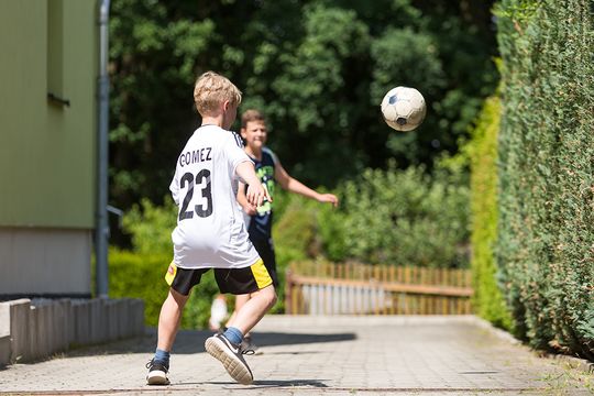 Zwei blonde Jungen spielen auf dem Hof ausgelassen Fußball. Der Ball fliegt gerade durch die Luft und die kleinen Fußballer versuchen den Ball als erstes zu treffen.