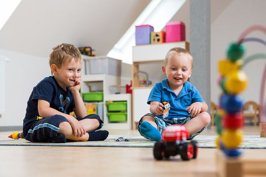 Im Spielzimmer des Suchthilfezentrums für Mutter und Kind spielt ein kleiner blonder Junge im blauen T-Shirt fröhlich mit einem roten Auto. Ein etwas älterer Junge sitzt im Schneidersitz daneben und lächelt verschmitzt.
