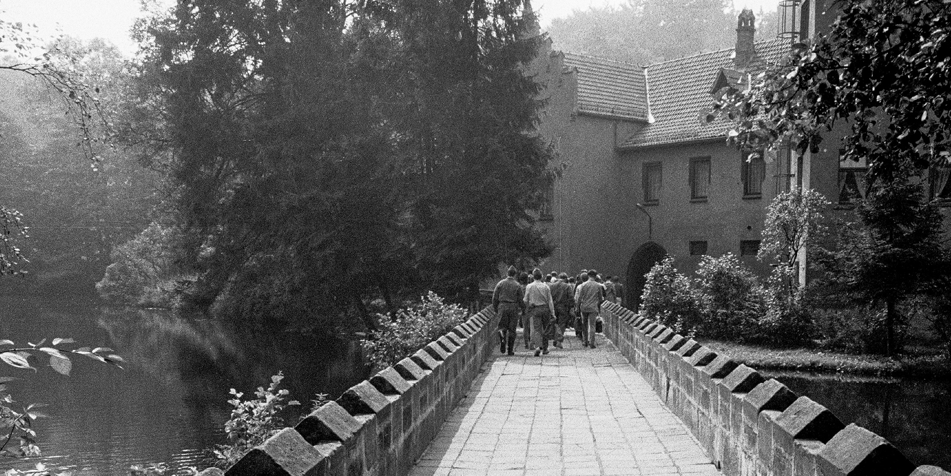 Schwarz-Weiss-Aufnahme: Blick auf Zöglinge des Jugendwerkhofs „Neues Leben“ die über Brücke zum Tor der Anlage laufen