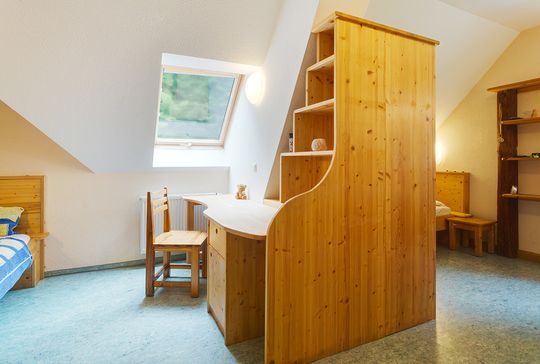 Ansicht eines Bewohnerzimmers. Ein großes Holzregal mit integriertem Schreibtisch trennt den Raum  in zwei Hälften, in denen sich jeweils ein Bett befindet.