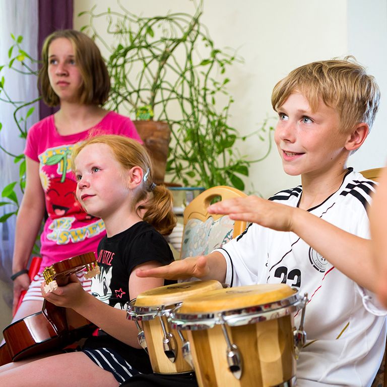 Nahaufnahme der Kinder im Kinderhaus in St. Gangloff beim Musikprojekt. Ein blonder Junge trommelt, das kleine Mädchen daneben spielt Gitarre.