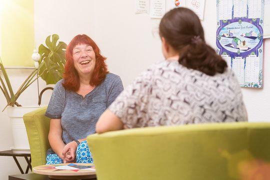 Eine jungen Mutter und eine symphatische Mitarbeiterin des Suchthilfezentrums für Mutter und Kind in apfelgrüne Sesseln im vertraulichen Gespräch. Die Mitarbeiterin schlägt die Hände zsammen und lacht herzlich.