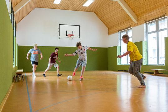 Vier Jungs in kurzen Hosen spielen in der kleinen Turnhalle des Jugendhilfezentrums Wolfersdorf Fußball. Der Junge in der Mitte des Bildes holt gerade mit seinem Bein aus, um den Ball seinem Gegenüber zuzuspielen.