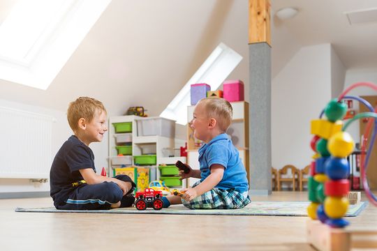 Zwei kleine blonde Jungen im Spielzimmer des Suchthilfezentrums für Mutter und Kind. Sie sitzen sich auf einem Spielteppich im Schneidersitz gegenüber und albern herum.  Um sie herum gibt es viel buntes Spielzeug.