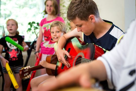 Nahaufnahme eines Jungen, der eine rote Gitarre auf den Knien hält und konzentriert spielt. Die Kinder im Hintergrund lauschen gespannt auf die Klänge.